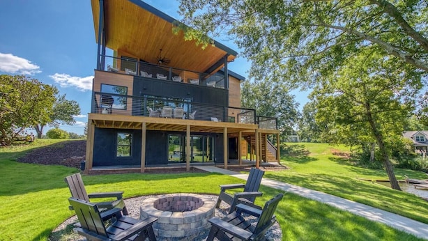 Een modern huis met twee verdiepingen, een overdekt terras, een vuurkorf en buitenmeubels.