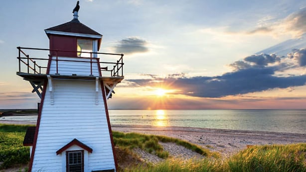 A white lighthouse with a red roof and balcony, situated on a grassy beach with the sun setting in the background.