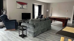 Una sala de estar con un sofá gris, un sillón negro, una mesa de billar y un televisor.