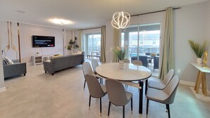 Ein modernes Wohnzimmer mit einem grauen Sofa, einem Esstisch mit Stühlen, einem an der Wand montierten Flachbildfernseher und einem großen Fenster mit Vorhängen.