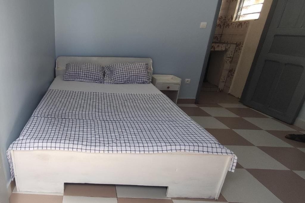 Una cama individual con colcha a cuadros, una mesita de noche y una ventana con vistas a una zona empavedada.