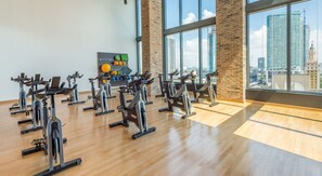 A gym with stationary bikes arranged in rows, a wall-mounted exercise board, and a view of the cityscape through large windows. - Miami Panorama Apartments (Miami)