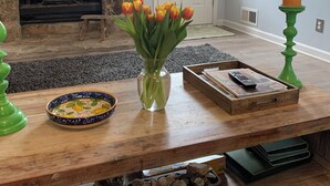 Una sala de estar con chimenea de piedra, un televisor montado arriba, una puerta con ventanal y una mesa con un jarrón de tulipanes, un cuenco y una bandeja con libros y un control remoto.