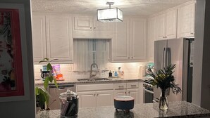 Una cocina con armarios blancos, encimera de granito, refrigerador, pileta y un florero con flores.