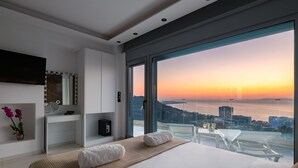 A modern bedroom with a large bed, a vanity with a mirror, and a view of the sunset over the water. - Rhodes Skyline Suite with out Door Jacuzzi Sea View A2 (Rhodes)
