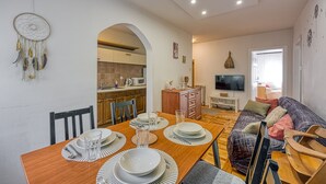 A dining area with a wooden table set for four, a kitchen in the background, and a living room with a sofa and TV.