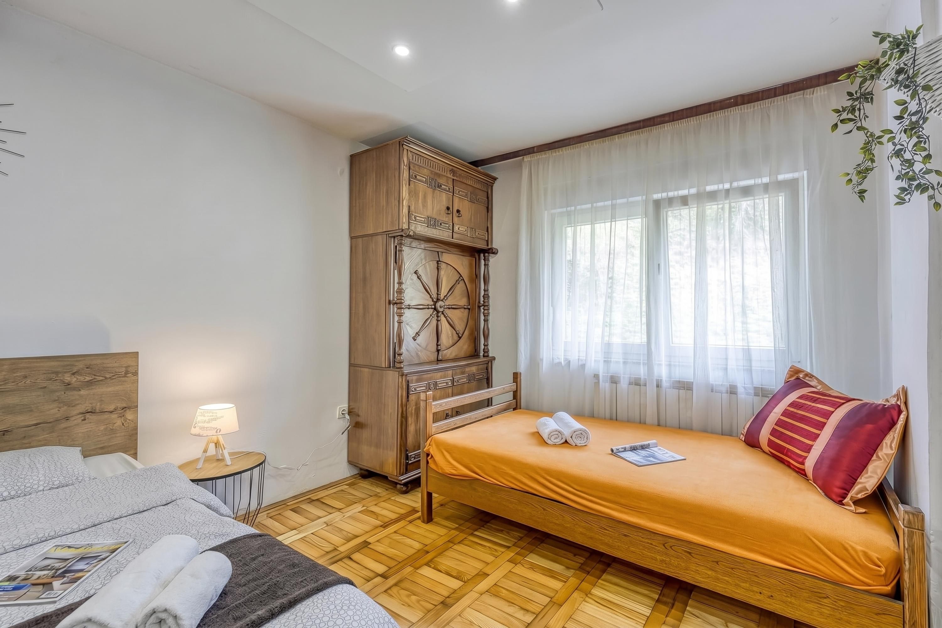 A hotel room with a bed, a wooden cabinet, a small table with a lamp, and a window with sheer curtains.