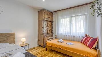 A hotel room with a bed, a wooden cabinet, a small table with a lamp, and a window with sheer curtains.