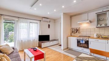 A modern living room with a flat-screen TV, a sofa, and a red table. The kitchen features white cabinets, a stove, and a microwave.