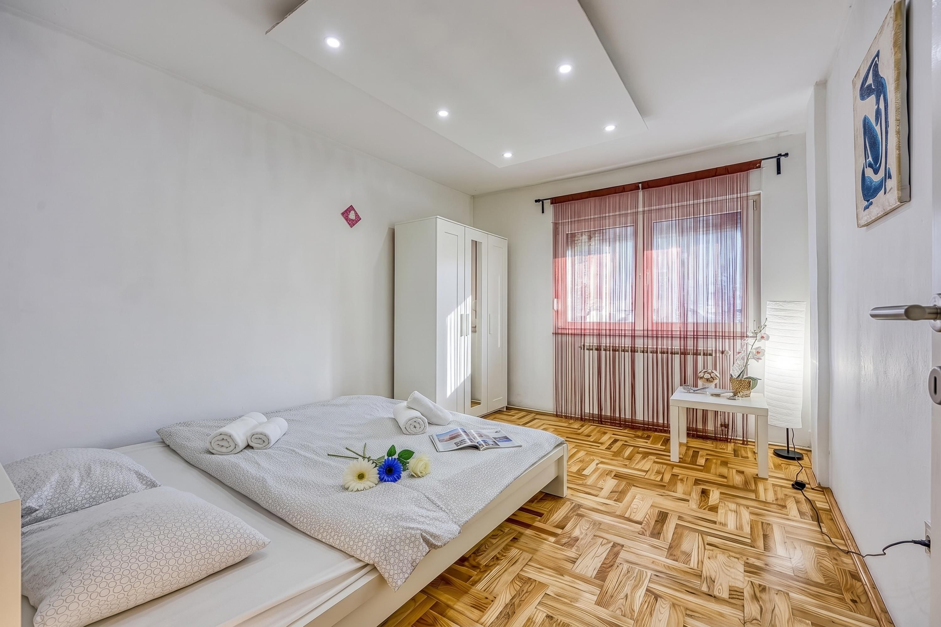 A hotel room with a bed, a bedside table, a window with curtains, and a wooden floor.