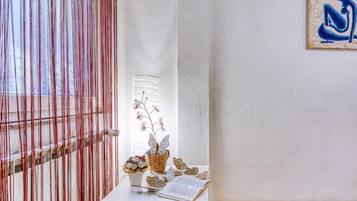 A room with a white wall, a wooden floor, a window with vertical blinds, a small table with a book, a vase with flowers, and a wall art piece.