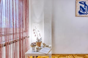 A room with a white wall, a wooden floor, a window with vertical blinds, a small table with a book, a vase with flowers, and a wall art piece. - Green Hill Zagreb - Apartment Sara, Zagreb, Croatia (Zagreb)