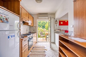 Een compacte keuken met houten kastjes, een inbouwfornuis en een koelkast. Er is een schuifpui die leidt naar een terras met tuinmeubilair.