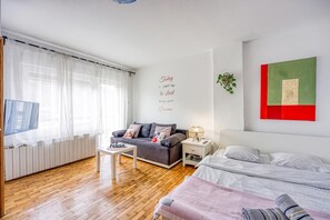 Un dormitorio moderno con un sofá gris, una cama con edredón blanco y gris con estampado, una mesita de noche blanca con lámpara, y una pared con una obra de arte verde y roja.