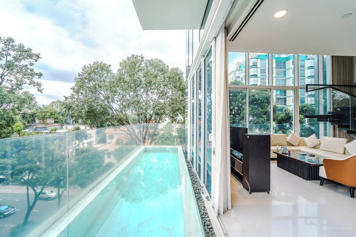 A modern living room with a glass balustrade overlooking a swimming pool, large windows, and a view of surrounding buildings.