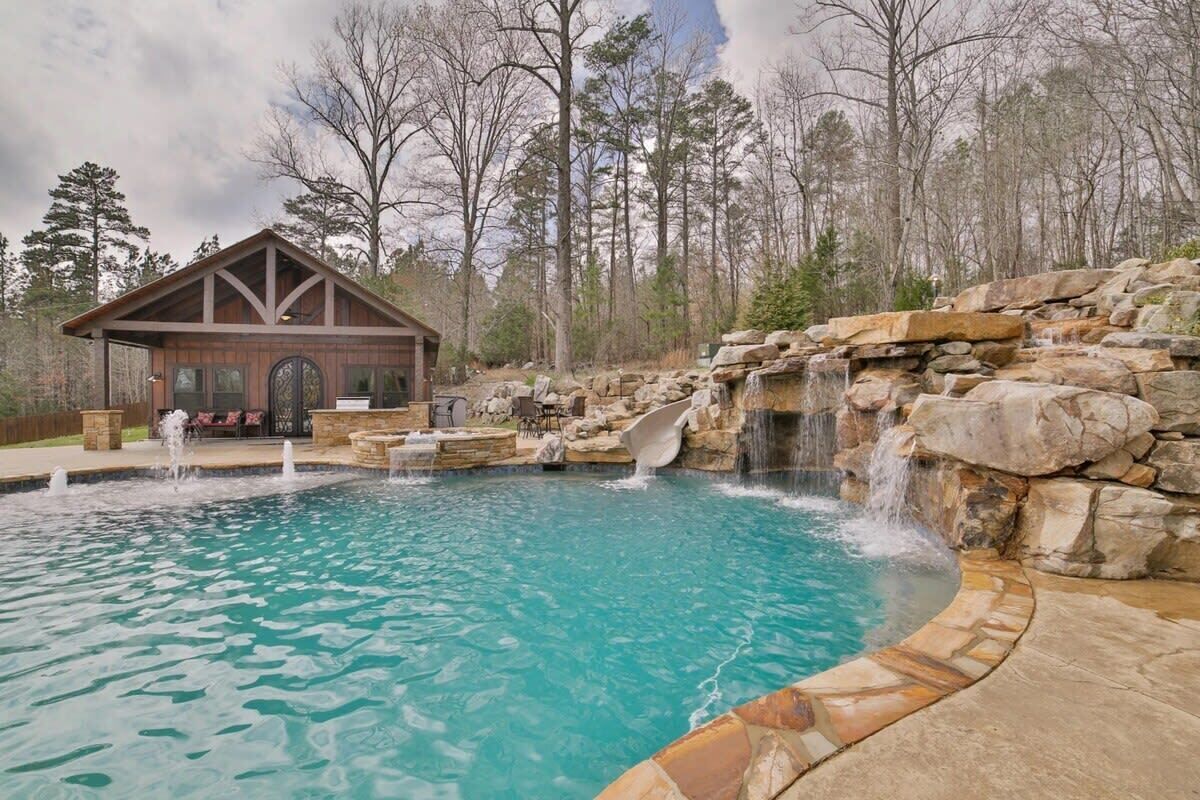 A swimming pool with a waterfall feature, surrounded by stone walls and a wooden building with a porch.