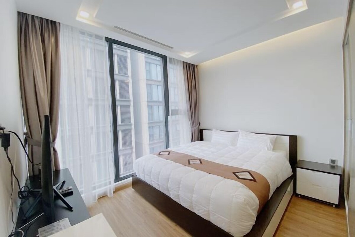 Quarto de hotel moderno com uma cama grande, uma escrivaninha com computador e uma janela com cortinas translúcidas.