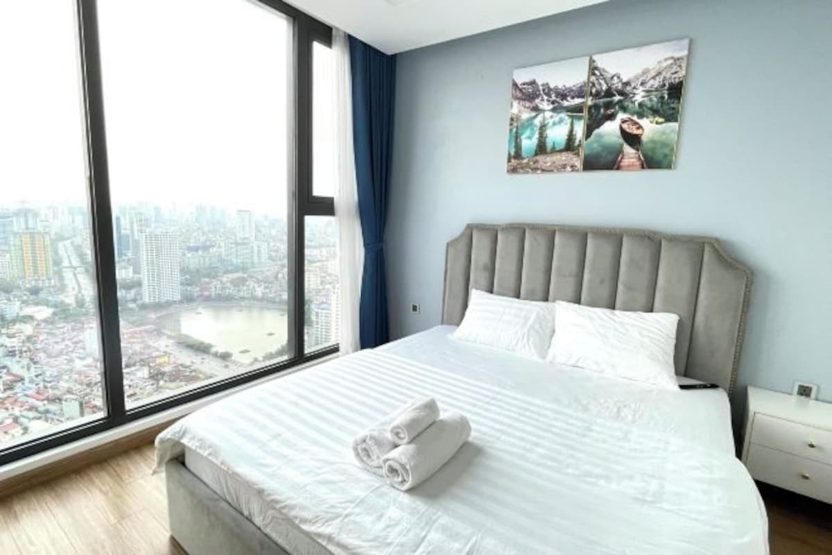 Quarto de hotel com uma janela grande, uma cama com roupa branca e vista para a cidade.