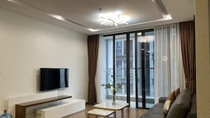 A modern living room with a flat-screen TV, a wooden coffee table, a sofa, and a large window with curtains. - Vinhomes Metropolis 3Brs  (Hanoi)