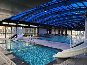 En indendørs swimmingpool med glasloft, vandrutsjebaner og liggestole.