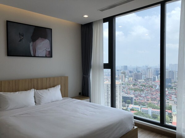 A hotel room with a large bed, a headboard, a painting on the wall, and a city view through a large window.