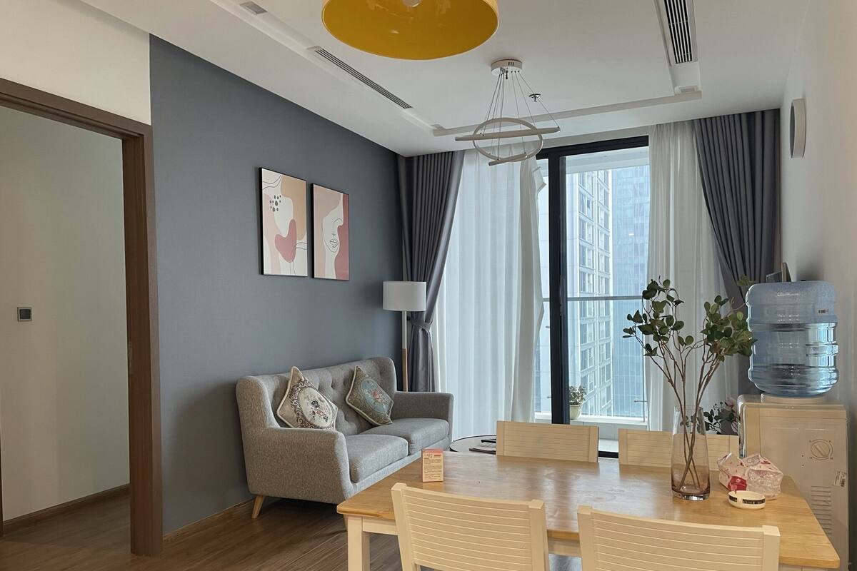 Ein modernes Wohnzimmer mit einem grauen Sofa, einem Holzespeisetisch mit vier Stühlen, einem Wasserspender und einem großen Fenster mit Vorhängen.