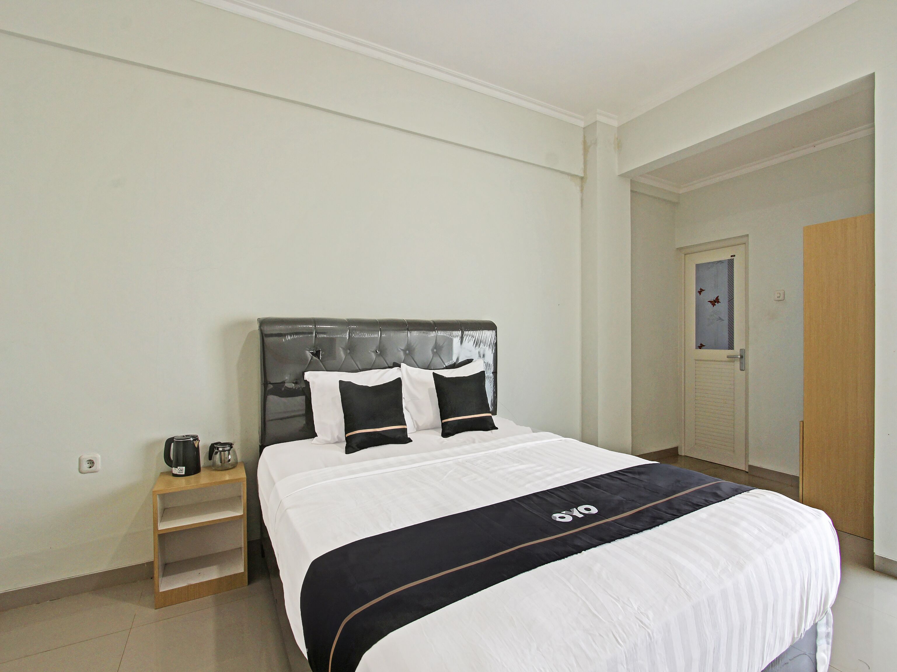 A neatly made bed with a dark headboard and white bedding, a bedside table with a kettle, and a door with a window view.
