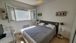 A bedroom with a bed, a desk with a laptop, a window with curtains, a bedside table with a lamp, and framed pictures on the wall. - 2-bedroom club villa with large terrace in holiday residence (SIX FOURS LES PLAGES)