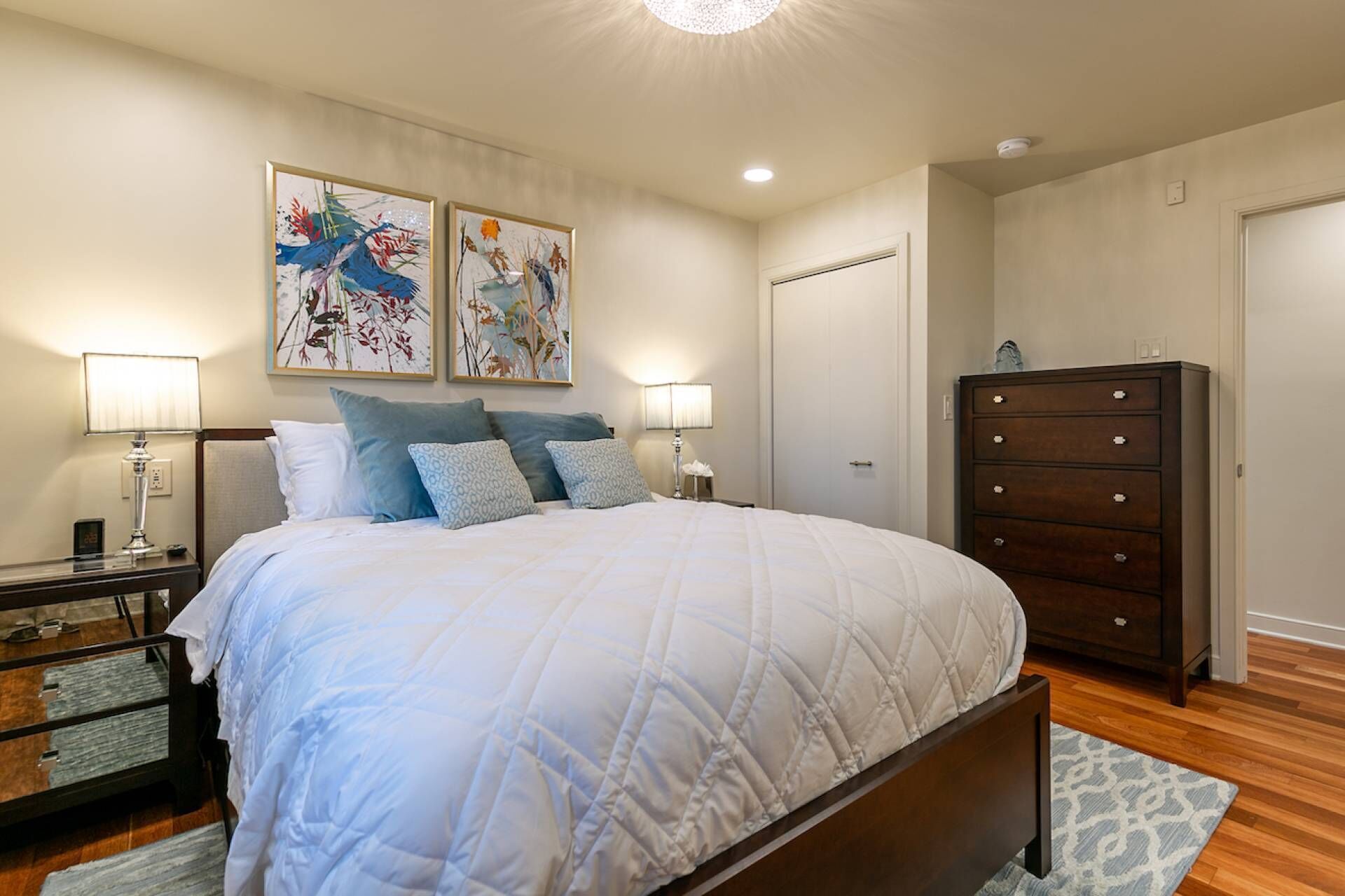 Une chambre à coucher comprenant un lit, deux tables de chevet avec des lampes, une commode et deux œuvres d’art encadrées accrochées au mur.