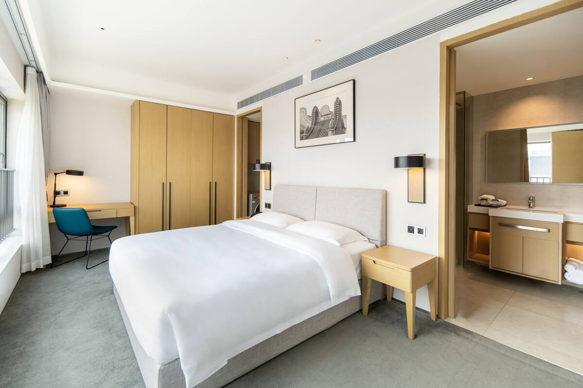 Une chambre d’hôtel avec un grand lit, un bureau avec une chaise, une armoire, une salle de bain avec un miroir et un lavabo, et un tableau encadré au mur.