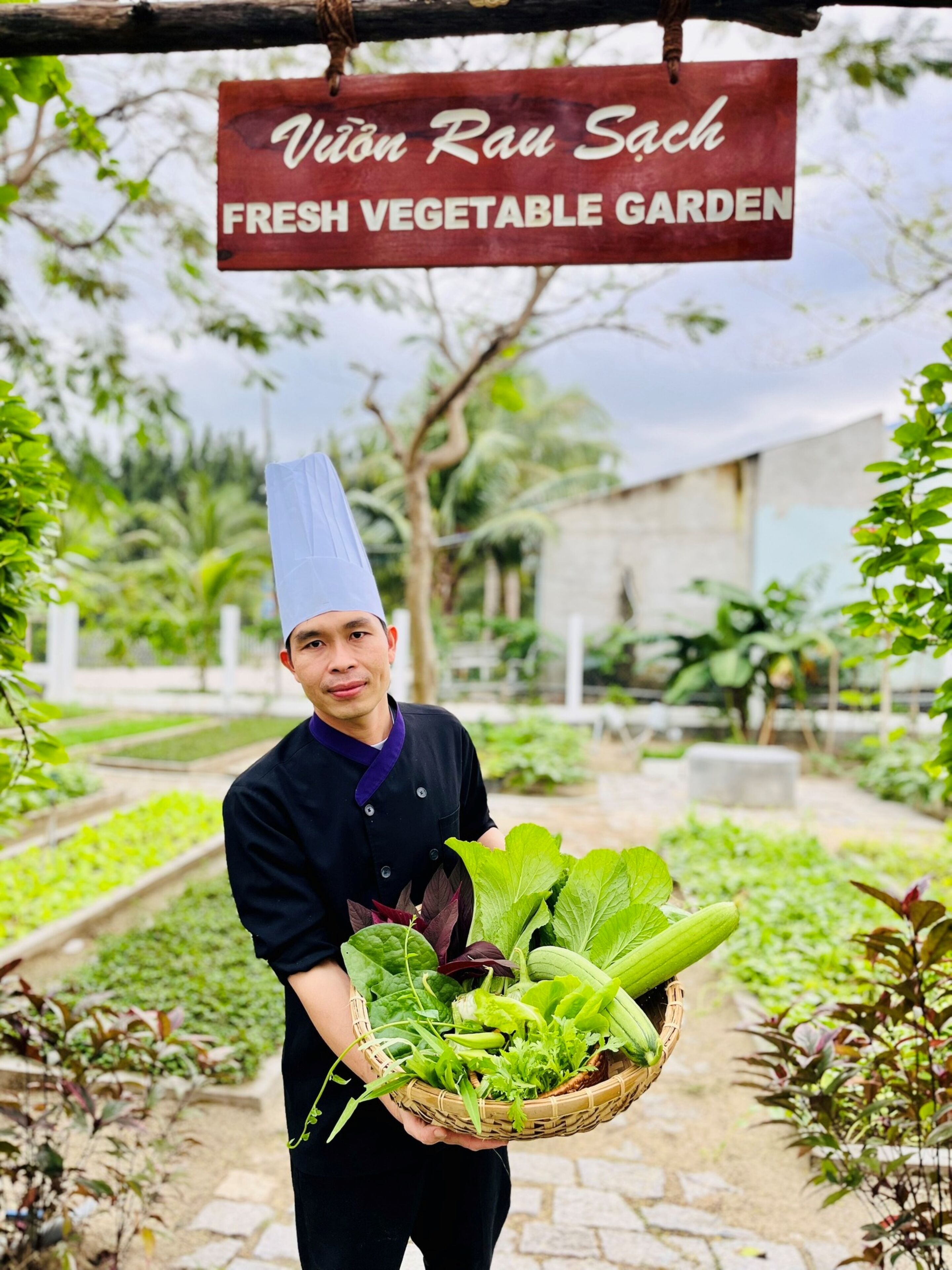 Un chef sostiene una cesta con verduras frescas frente a un letrero que dice 'Vương Rau Sach Fresh Vegetable Garden'.