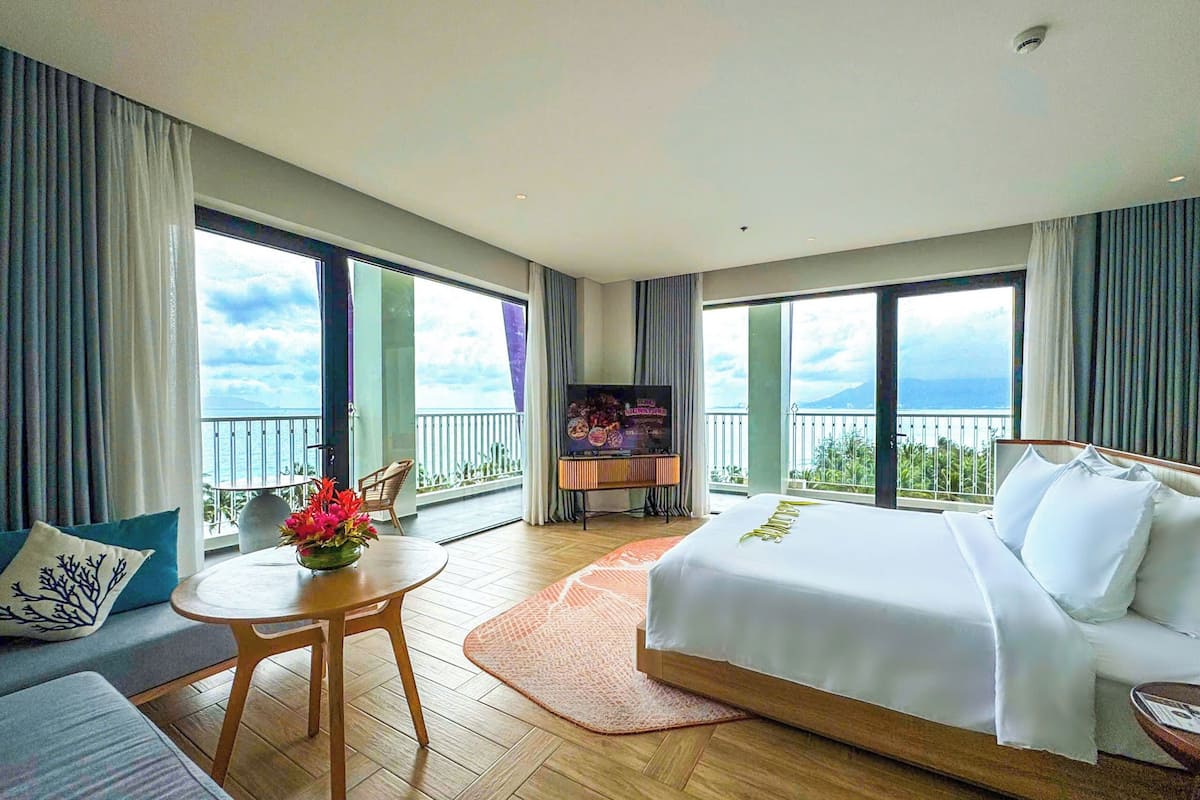 A hotel room with a bed, a TV, a table with flowers, and a balcony with a view.