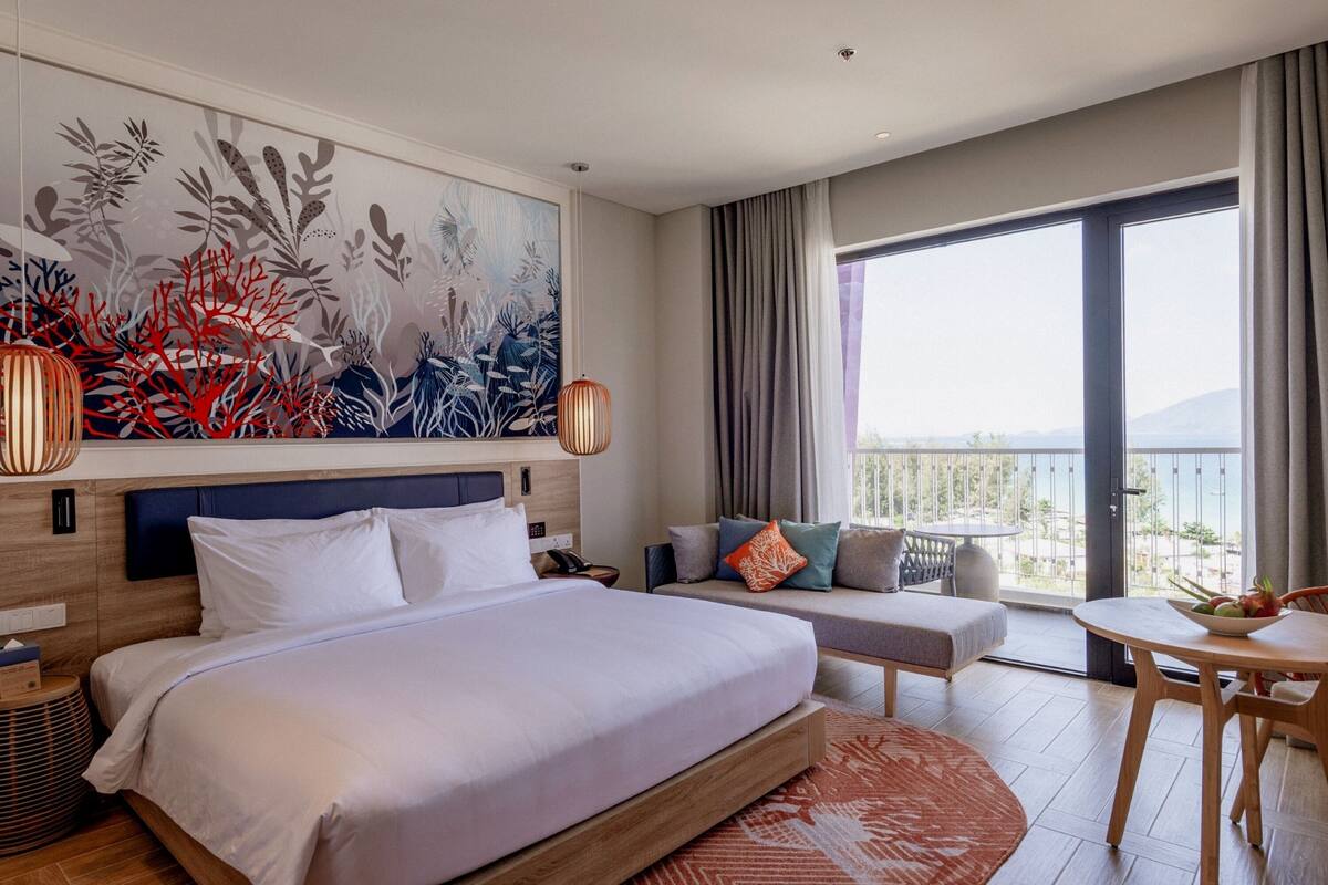 A hotel room with a large bed, a sofa, a dining table, and a balcony with a view.