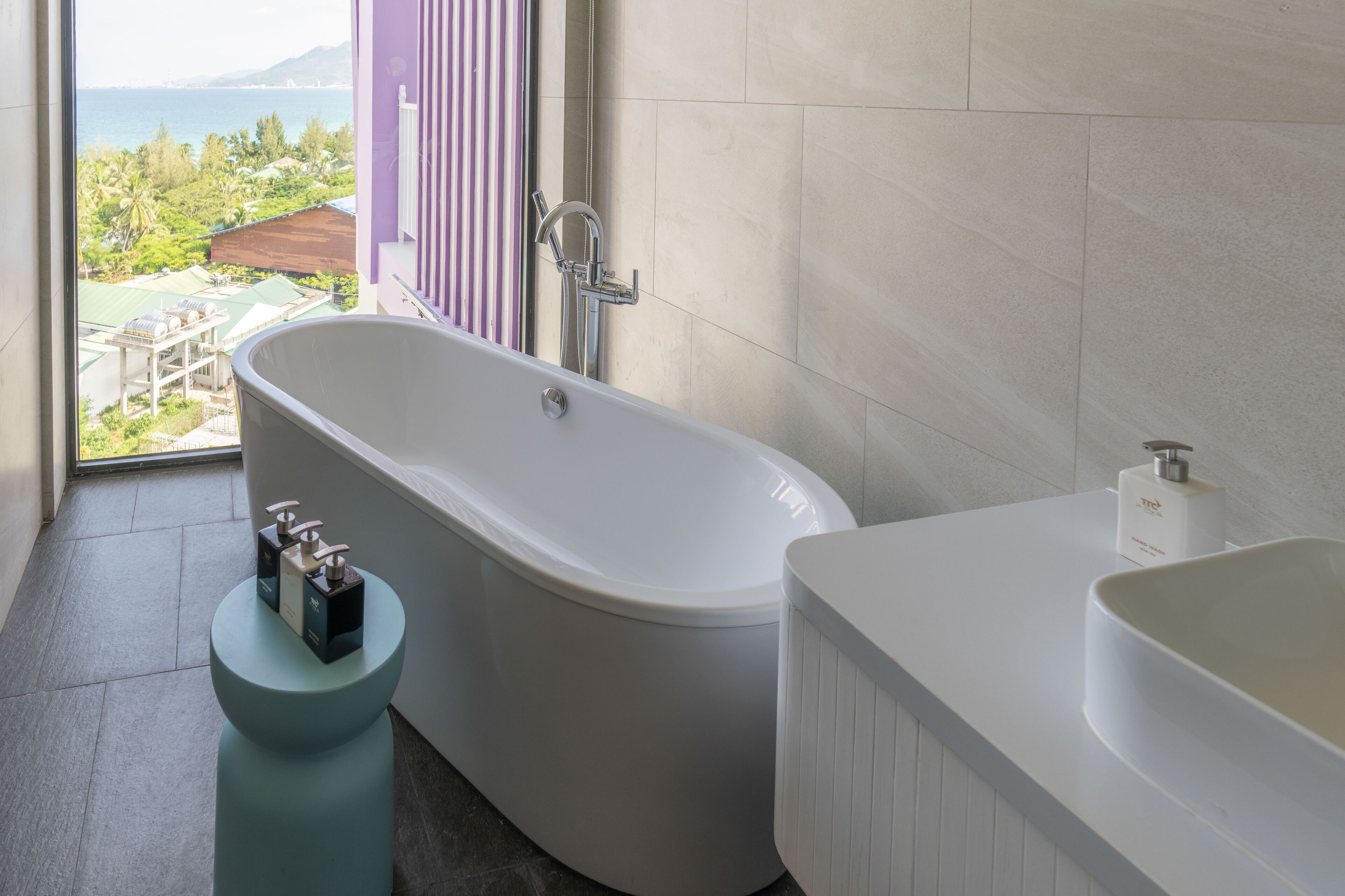 Une salle de bain moderne avec une baignoire sur pieds, un lavabo blanc et une vue sur un paysage tropical grâce à une grande fenêtre.