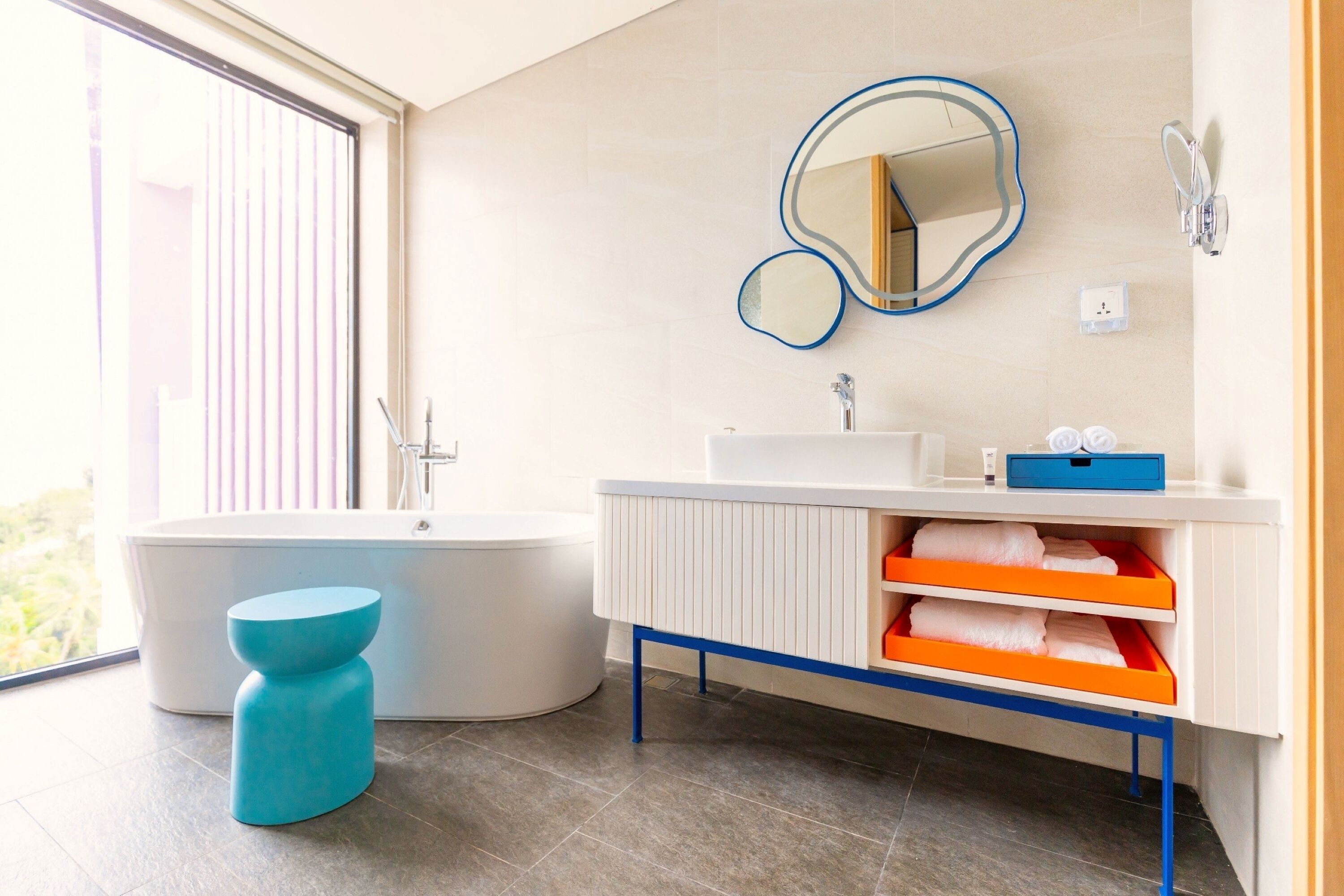 Une salle de bain moderne avec une baignoire, un meuble-lavabo blanc doté de rangements orange, un tabouret bleu et un grand miroir.