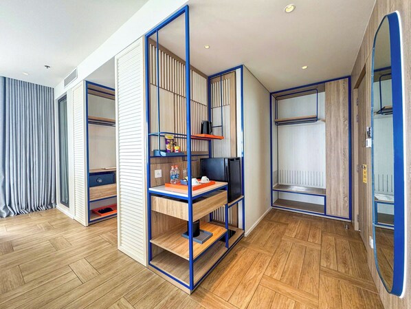 A modern hotel room with a wooden floor, a desk with a computer, a chair, a wardrobe, and a mirror.