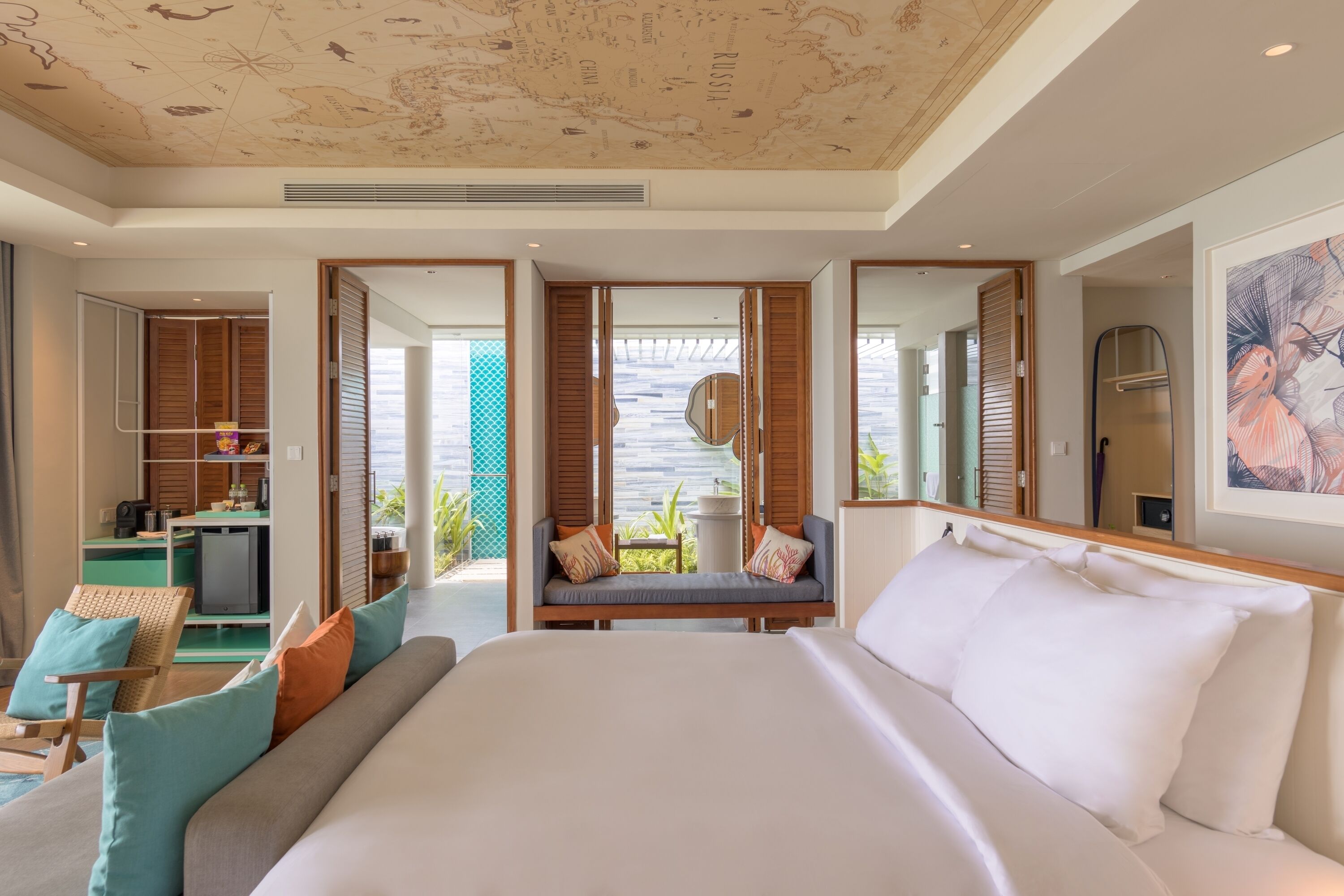 A modern hotel room with a large bed, a seating area with cushions, a kitchenette, and a view of the outdoors.