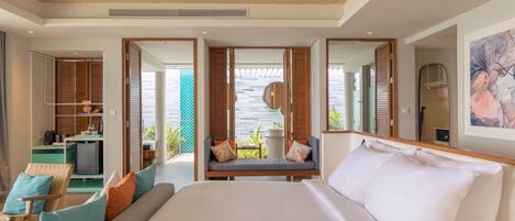 A modern hotel room with a large bed, a seating area with cushions, a kitchenette, and a view of the outdoors.