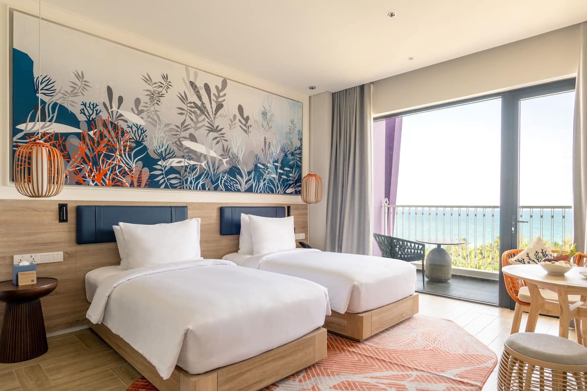 A hotel room with two beds, a large window with a view, a painting on the wall, and a balcony with a table and chairs.