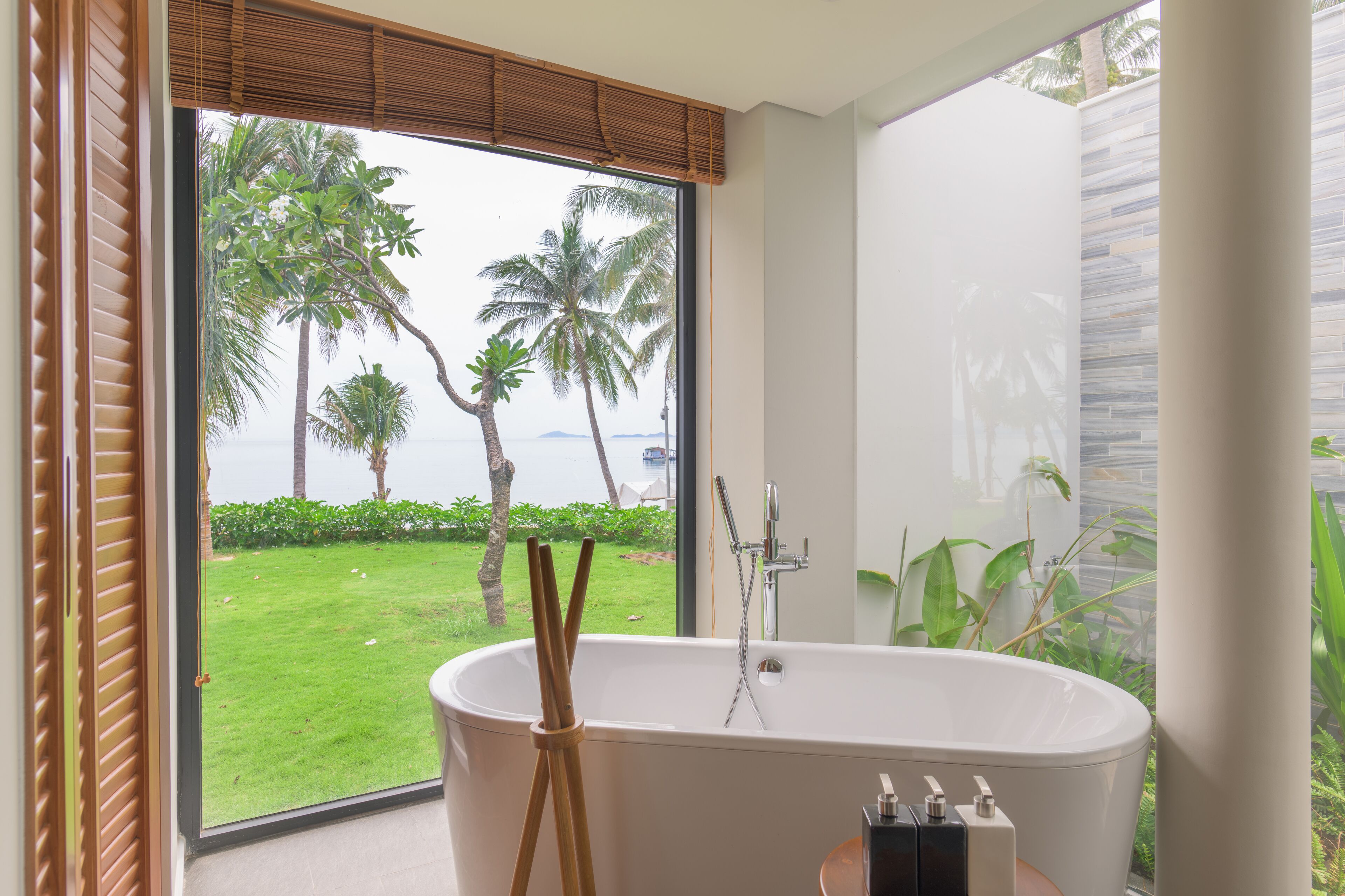 Een moderne badkamer met een vrijstaand bad, een groot raam met uitzicht op een gazon en palmbomen, en een stenen muur.
