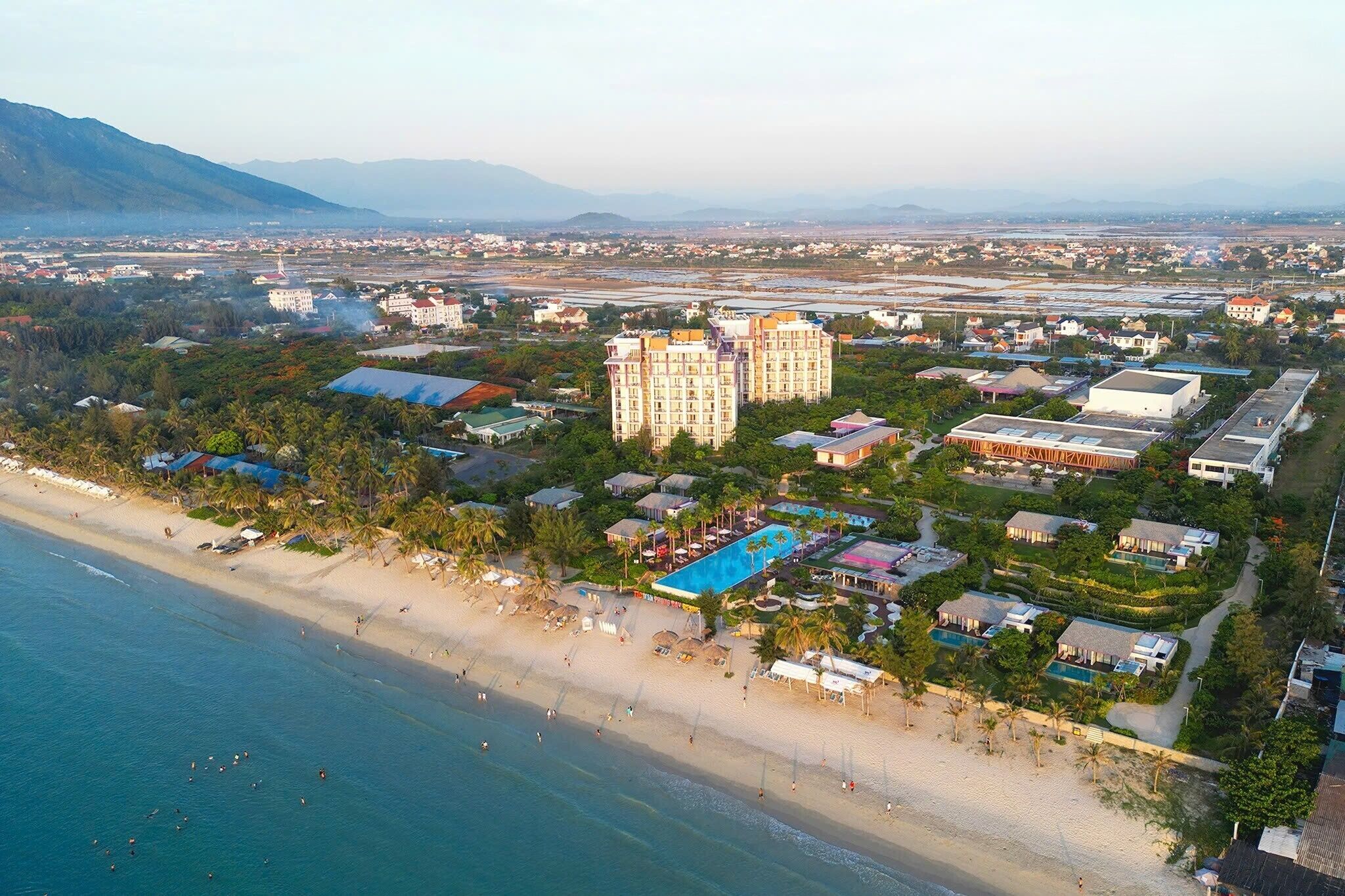 An aerial view of a coastal resort with a beach, a large swimming pool, and multiple buildings.