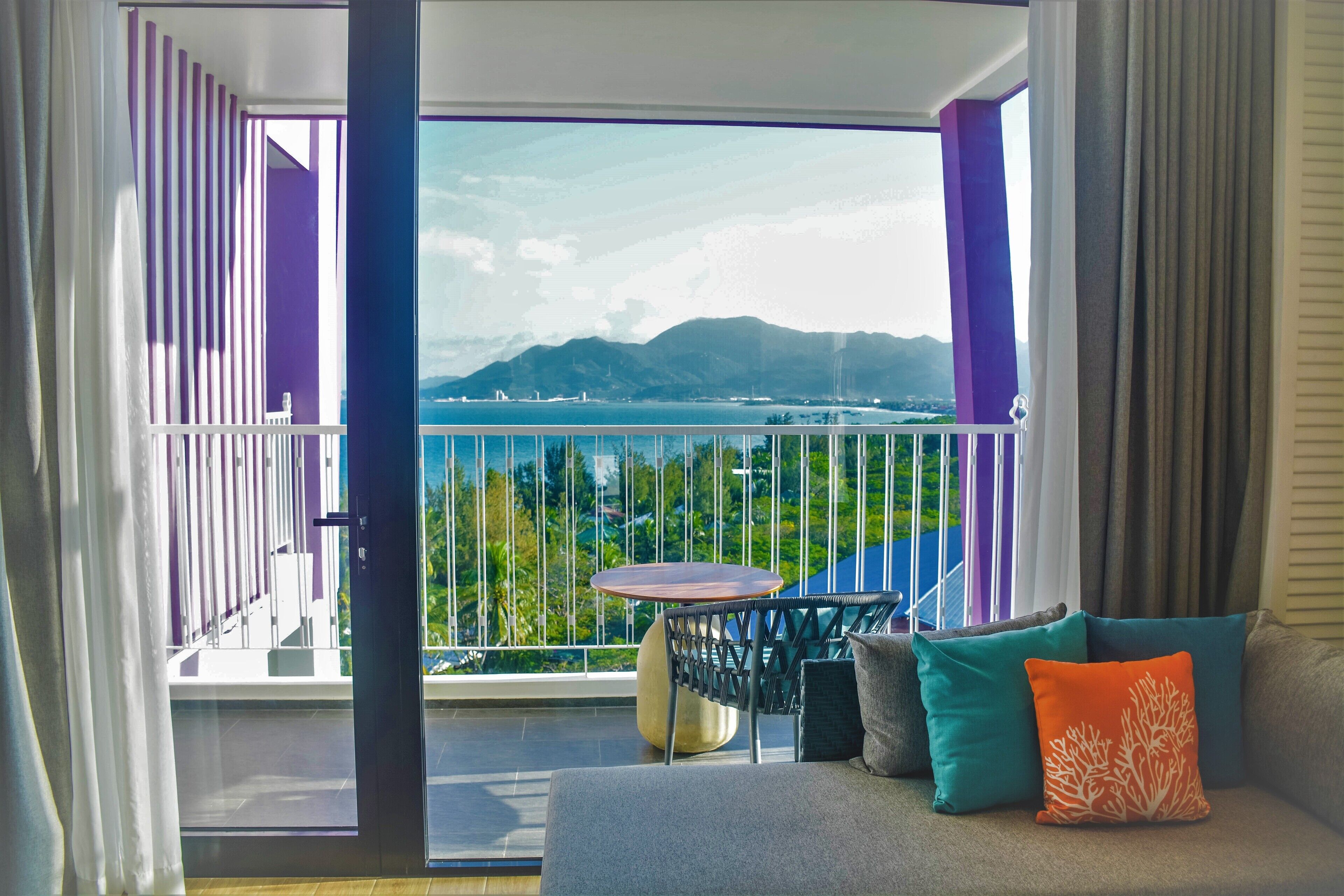 A balcony with a view of a body of water and mountains, a round table, and a sofa with cushions.