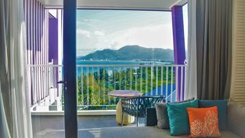 A balcony with a view of a body of water and mountains, a round table, and a sofa with cushions.