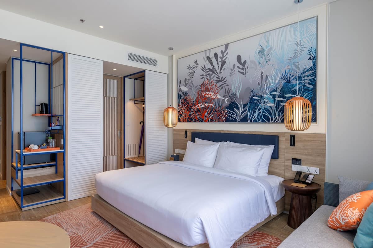 A modern hotel room with a large bed, a side table with a lamp, a wardrobe with a blue frame, and a large wall painting.