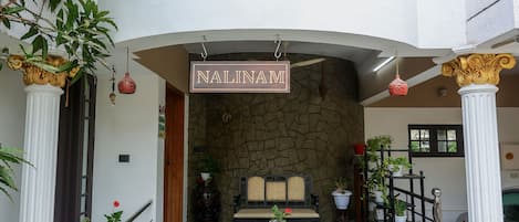 A hotel entrance with a sign reading "NALINAM HOME STAY," a stone facade, a wooden bench, and potted plants.