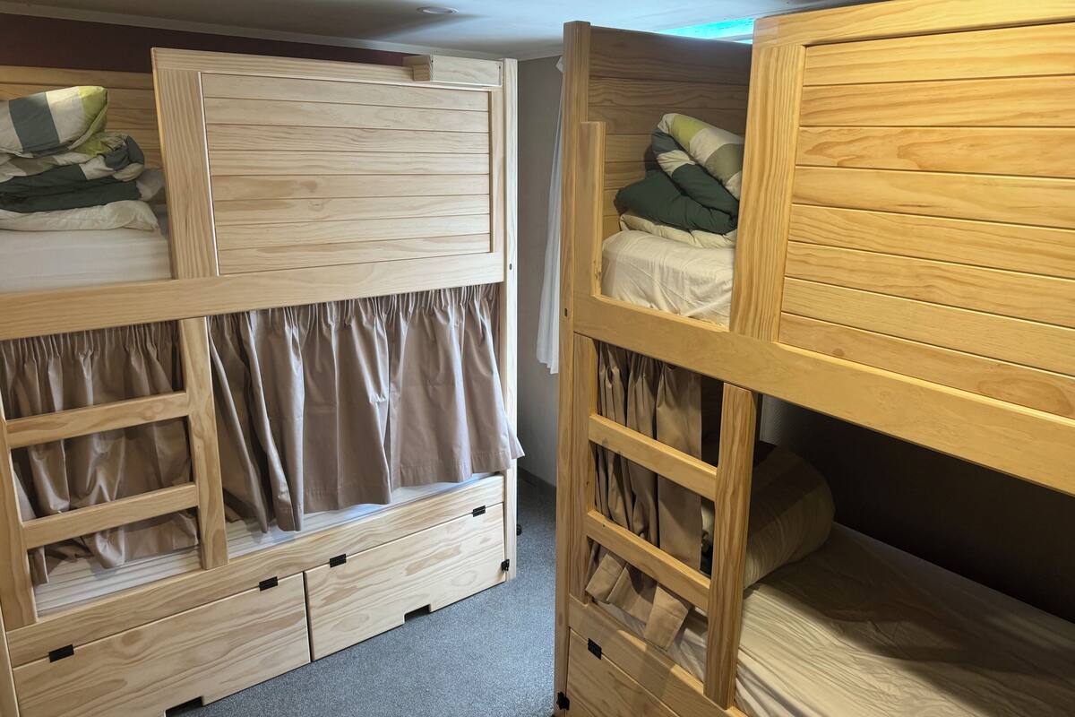 Une chambre avec des lits superposés dotés de têtes de lit en bois, une armoire intégrée avec des tiroirs et une fenêtre recouverte d’un rideau.