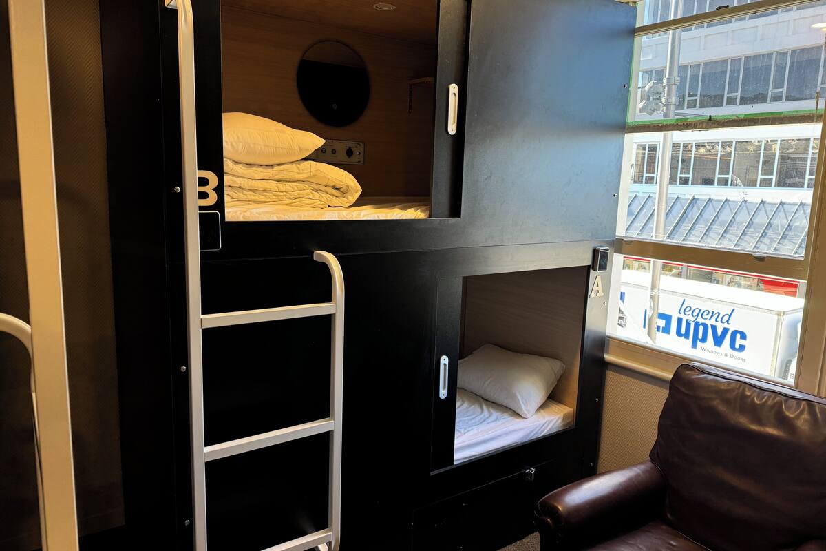 Une chambre avec un lit superposé, une échelle, une fenêtre donnant sur des immeubles et un fauteuil en cuir marron.