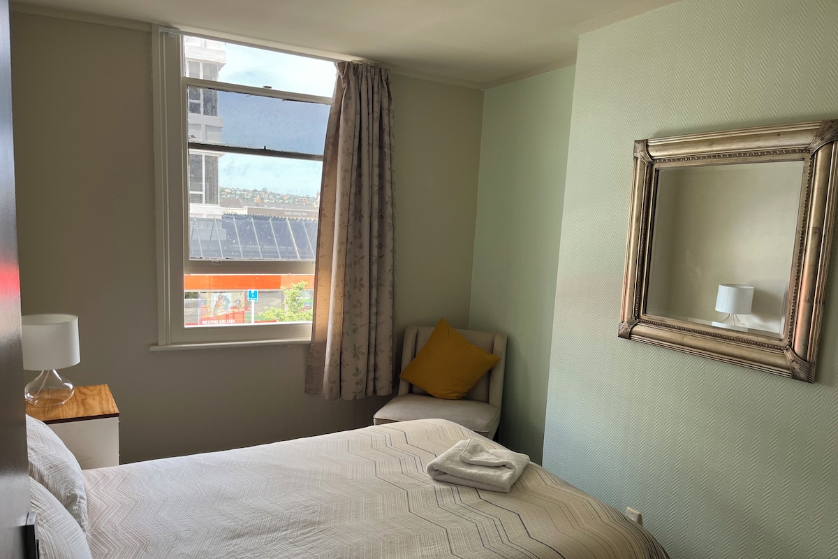 A hotel room with a bed, a chair, a window with curtains, a wall-mounted mirror, and a lamp on a bedside table.