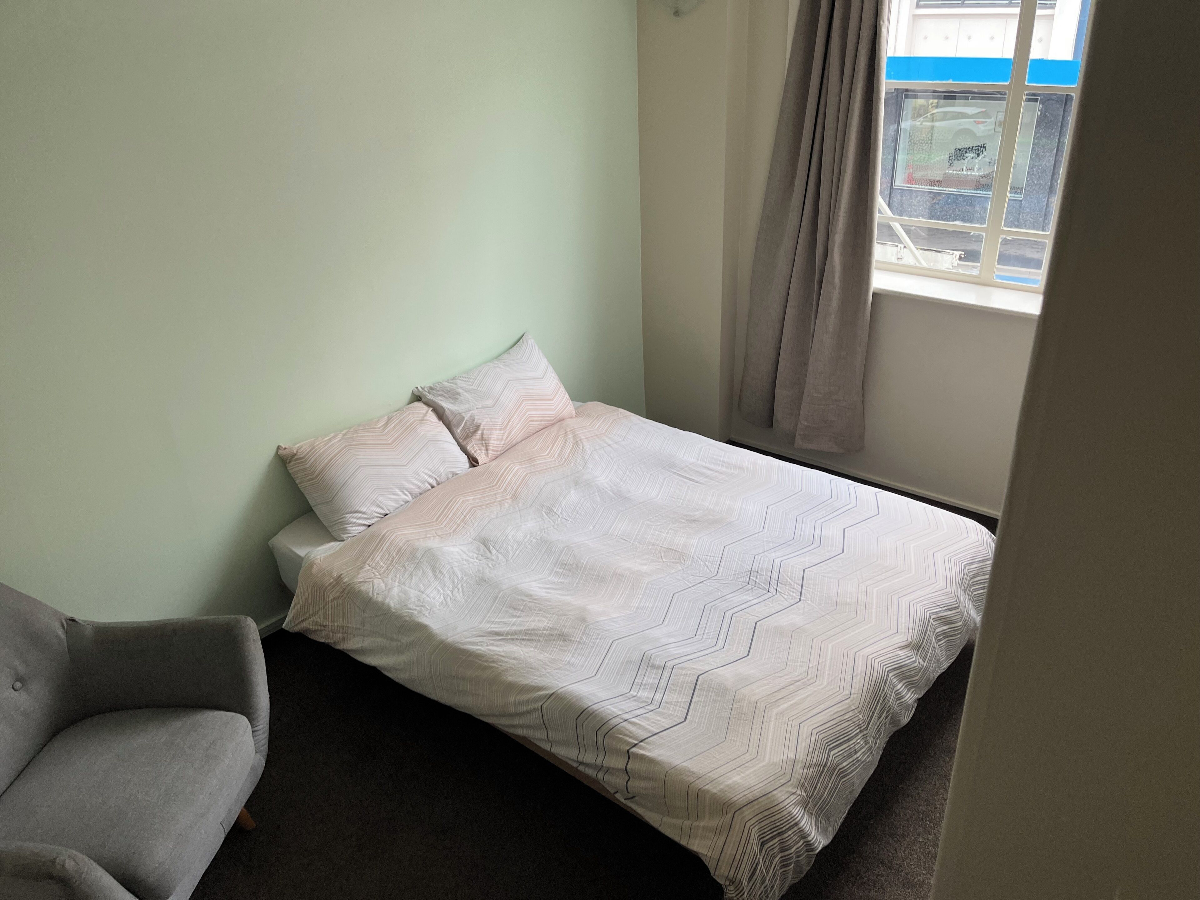 A small, neatly arranged bedroom with a single bed, a grey sofa, and a window with curtains.