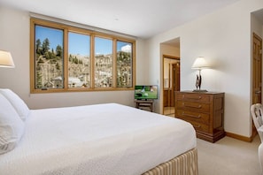 A bedroom with a large bed, a television, a dresser, and a window with a view of a mountainous landscape. - Highlands Lodge Beaver Creek - Corner Unit (Beaver Creek)